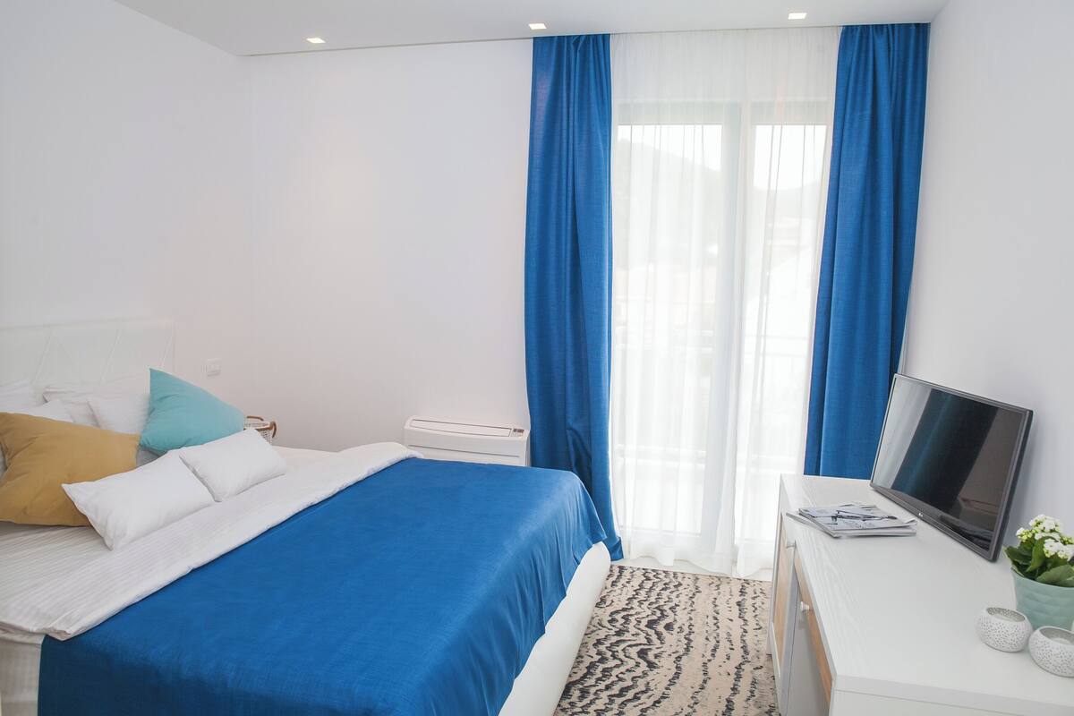 A hotel room with a bed, a desk with a laptop, a TV, and a window with blue curtains.