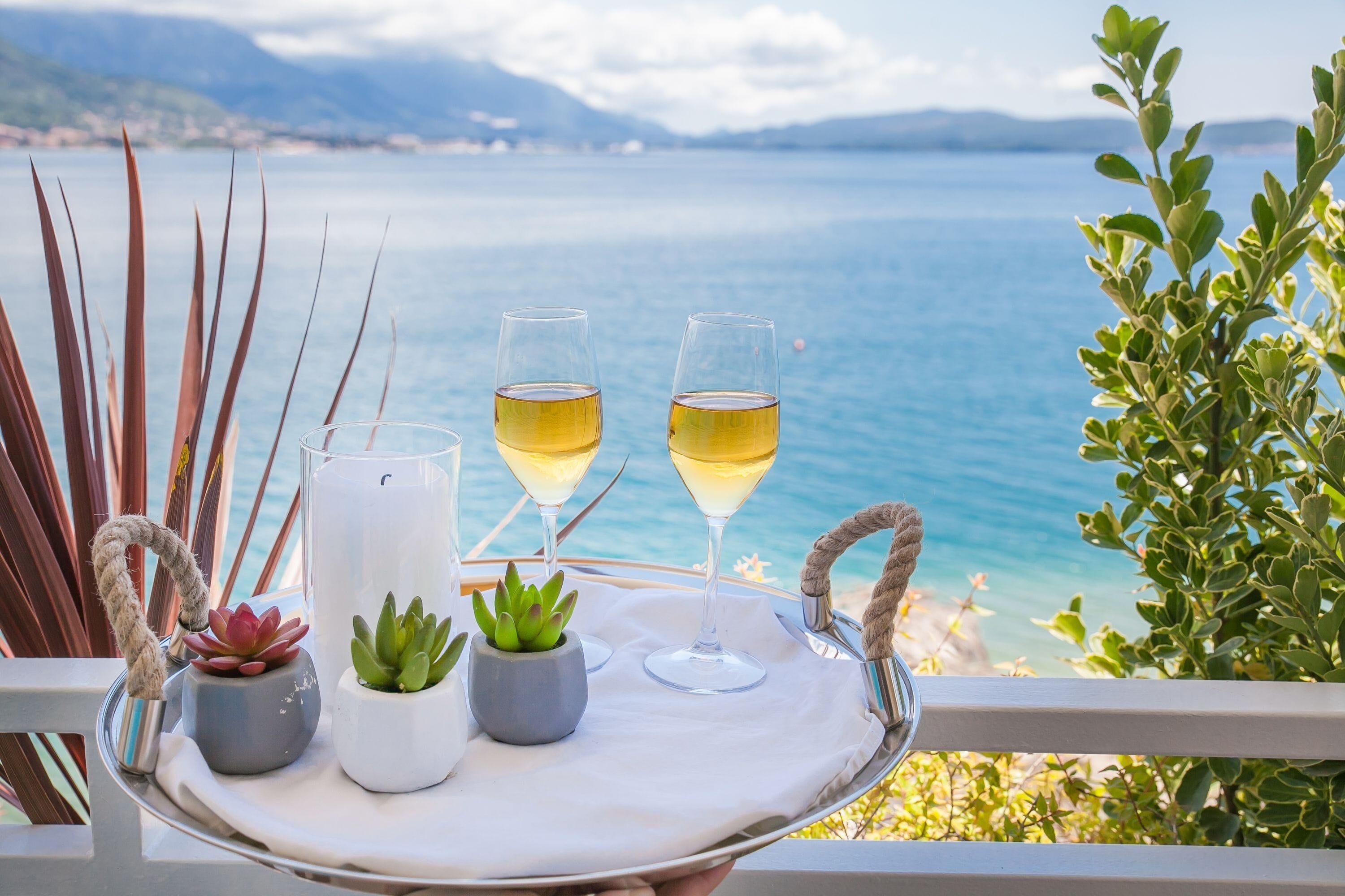 Un plateau avec deux verres de vin blanc, une bougie et de petites plantes en pot, posé sur un balcon donnant sur une étendue d’eau.