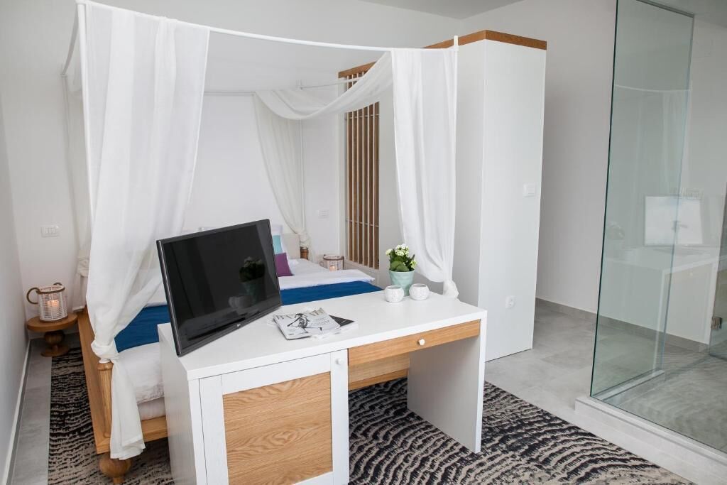 A bedroom with a bed, a desk with a laptop, a bedside table with a lamp, and a glass shower enclosure.