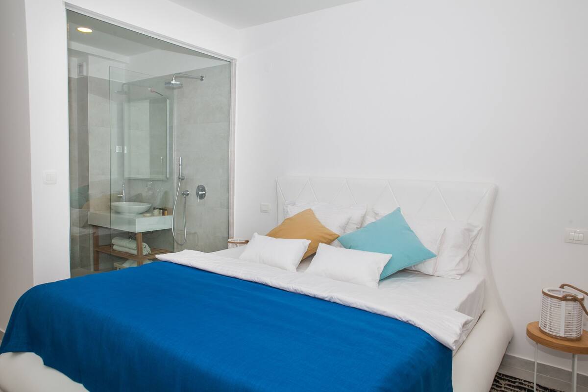 A modern bedroom with a large bed, a glass shower enclosure, and a white headboard.