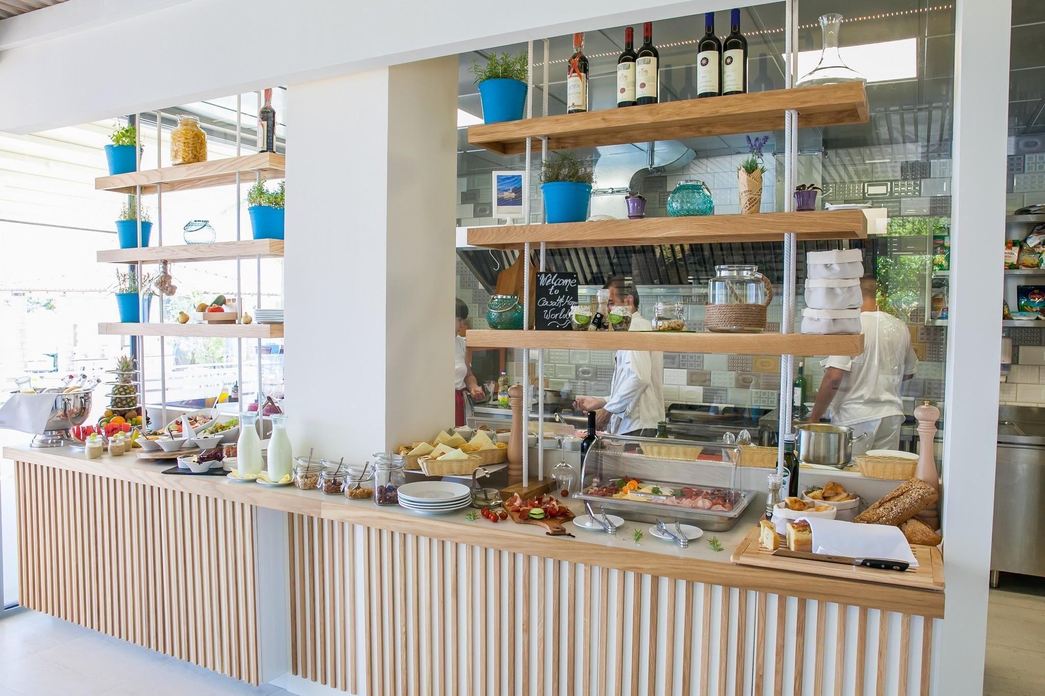 Ein Buffet mit verschiedenen Speisen, einer Weinauswahl und einem durch eine Glasscheibe einsehbarer Küchenbereich.