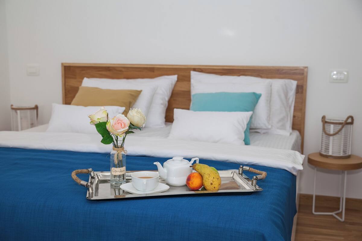 A neatly made bed with a blue bedspread, a tray with a teapot, cup, and fruit, and a vase with flowers.