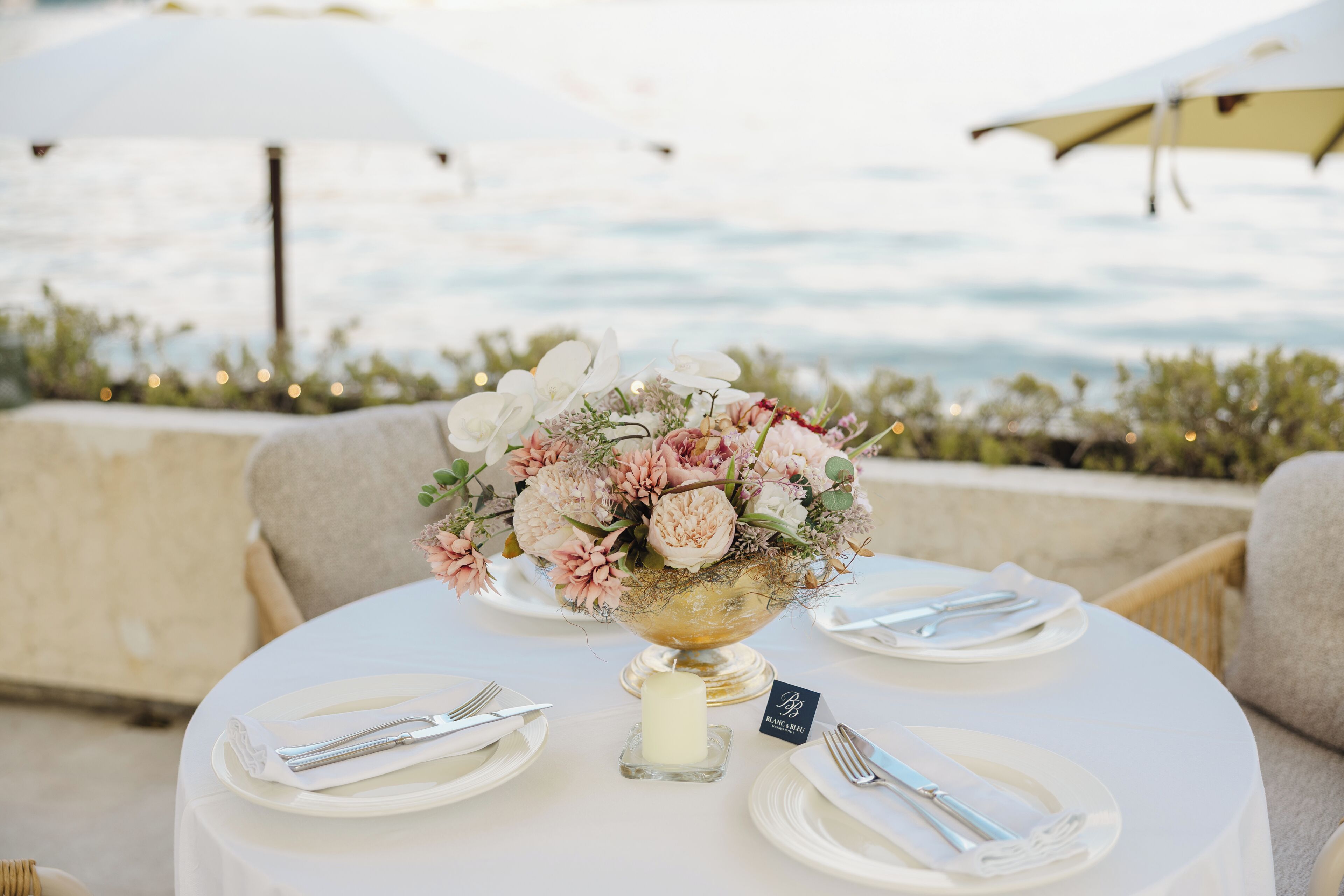 A round table with a floral centerpiece, white plates, cutlery, and a candle, set against a backdrop of a scenic view.