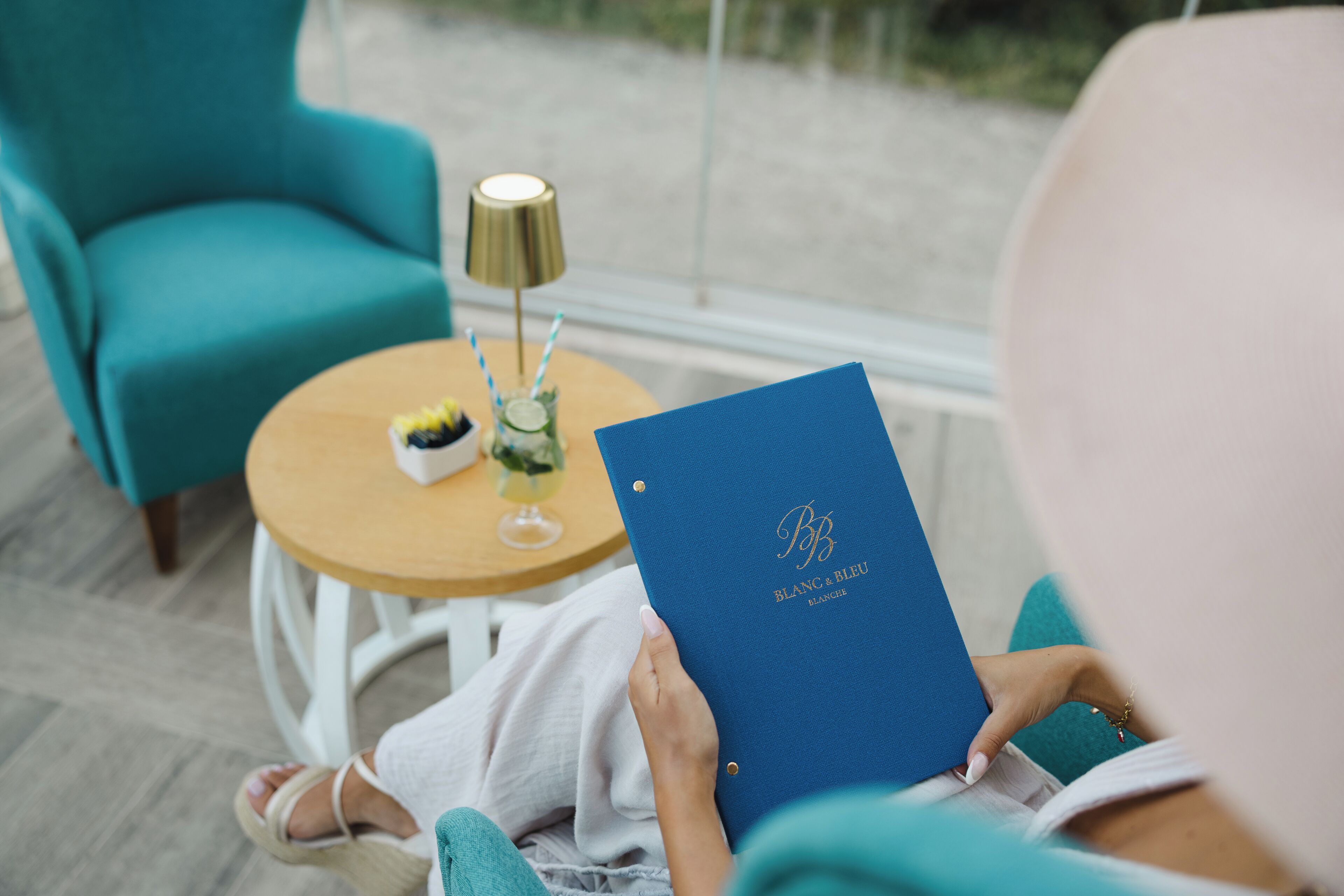 Une personne assise sur une chaise, avec un dossier bleu à portée de main, une petite table sur laquelle se trouve une lampe, et un verre avec une paille.