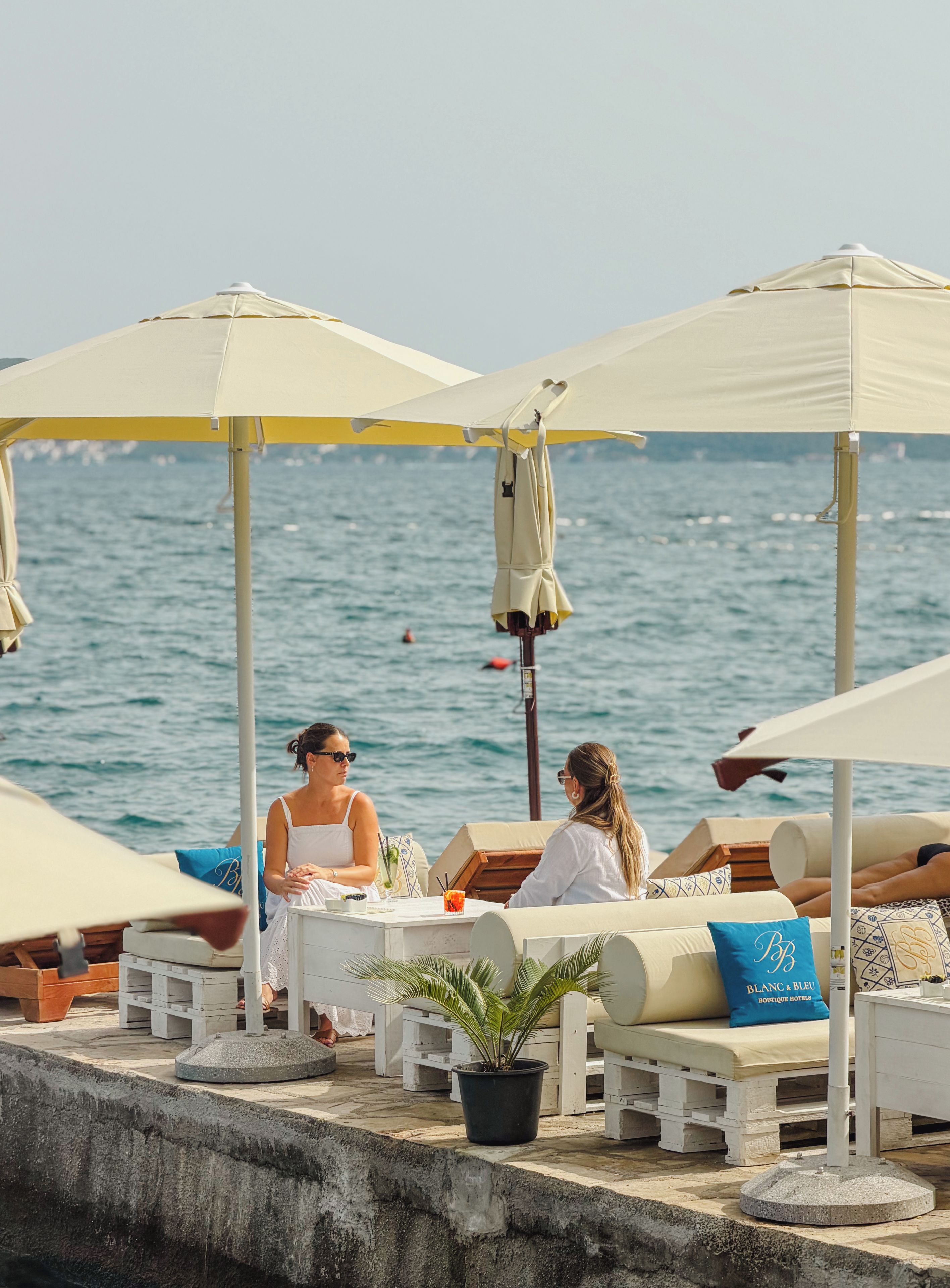 Een terras aan zee met buitenmeubilair en parasols, een plant in pot, en uitzicht op de zee.