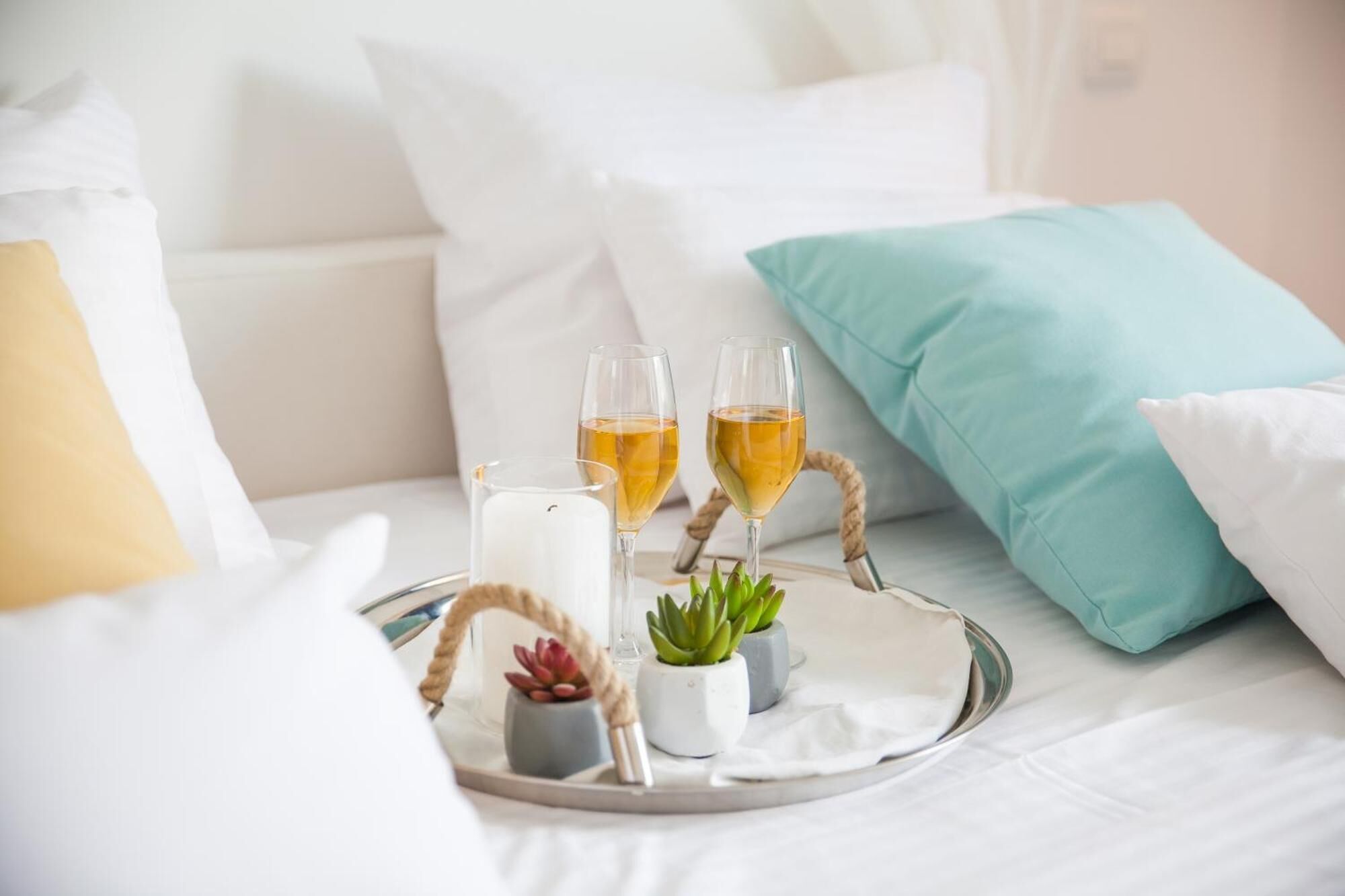 A tray with two glasses of white wine, a candle, and small potted plants on a bed with white bedding and teal pillows.
