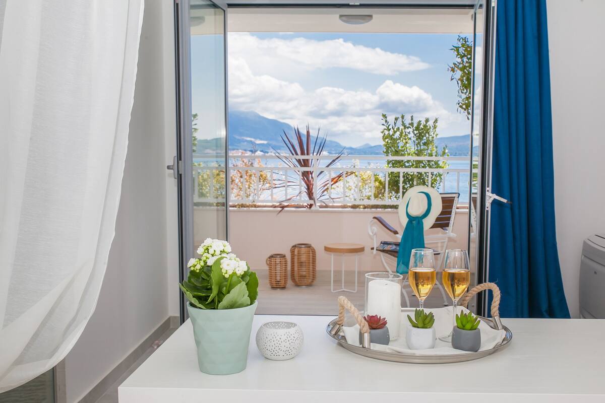 A balcony with a view of the sea, a potted plant, candles, and a tray with glasses and a book.