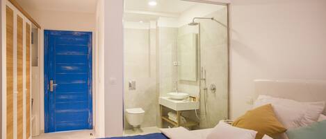 Un dormitorio con cama, una puerta azul, un baño con cabina de ducha de vidrio y un lavabo blanco.