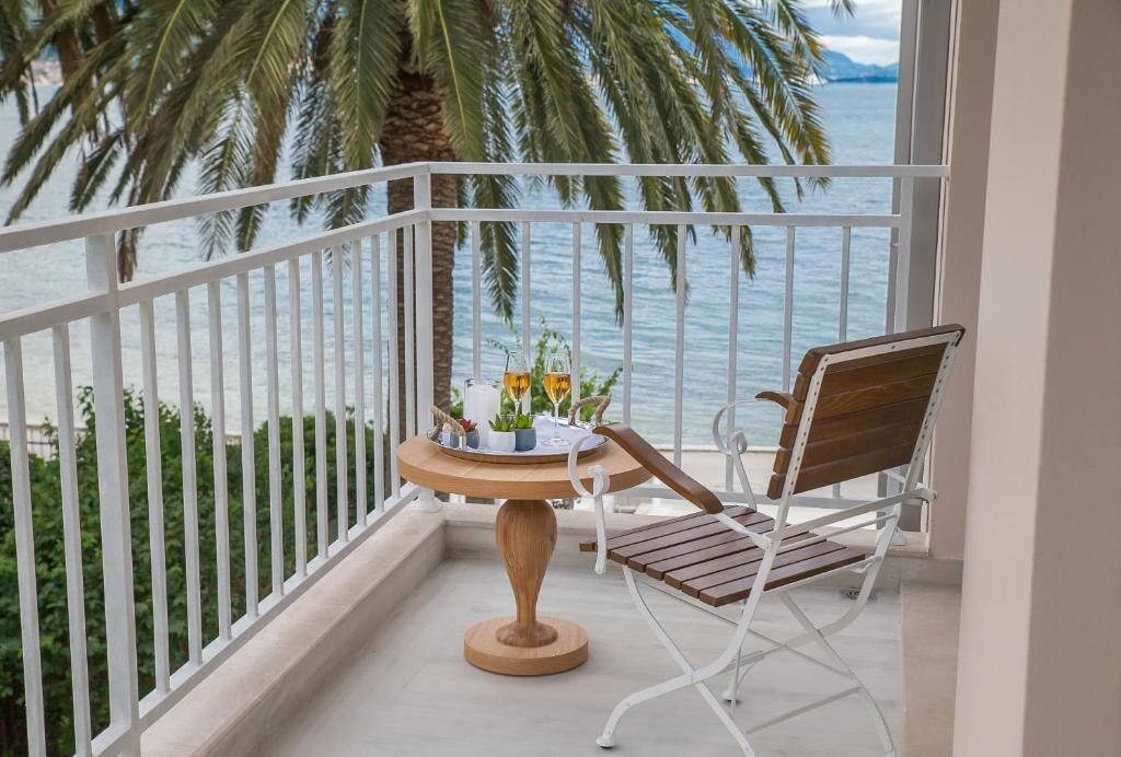 A balcony with a wooden table and two chairs, overlooking a body of water and palm trees.
