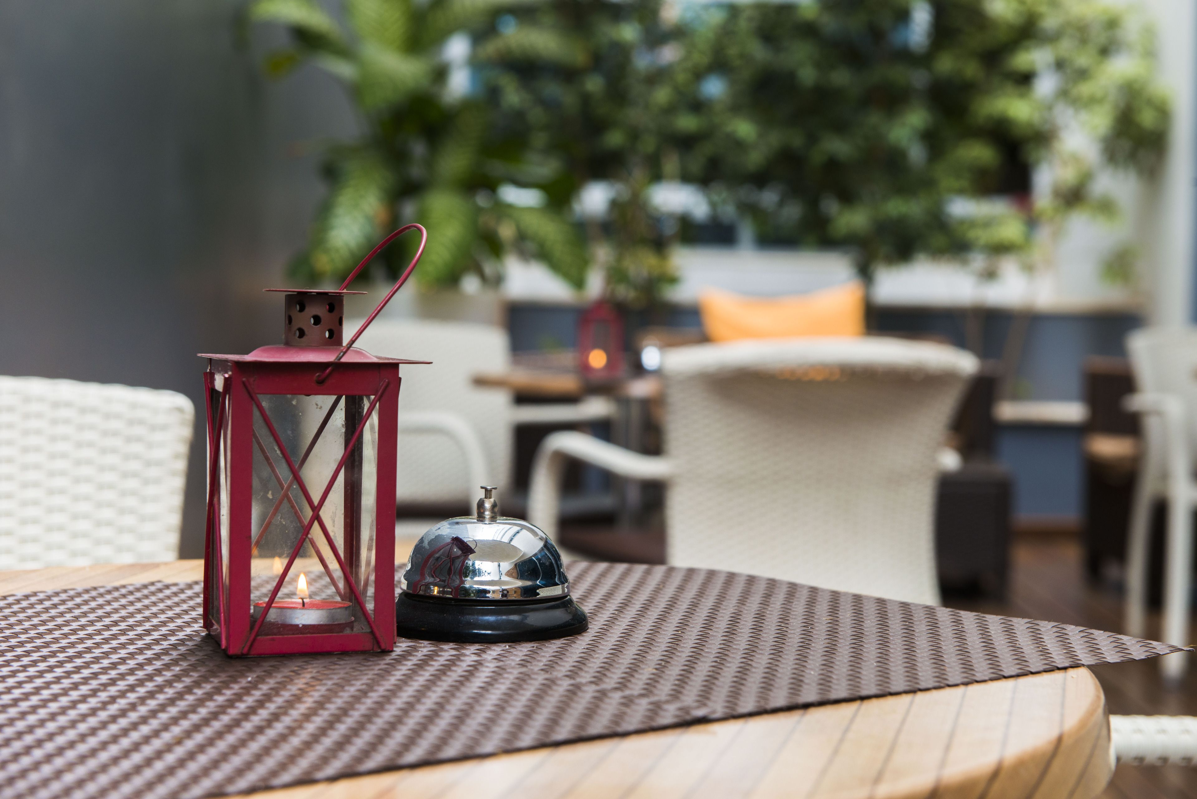 A table with a red lantern and a bell on a woven placemat, set in an outdoor dining area with wicker furniture and a view of greenery.