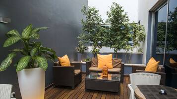 Un área de estar moderna al aire libre con mobiliario de mimbre, una planta grande en maceta y una mesa de vidrio.