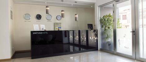 Zona de recepción de hotel con un diseño moderno, que incluye un mostrador de recepción, relojes y una planta en maceta.
