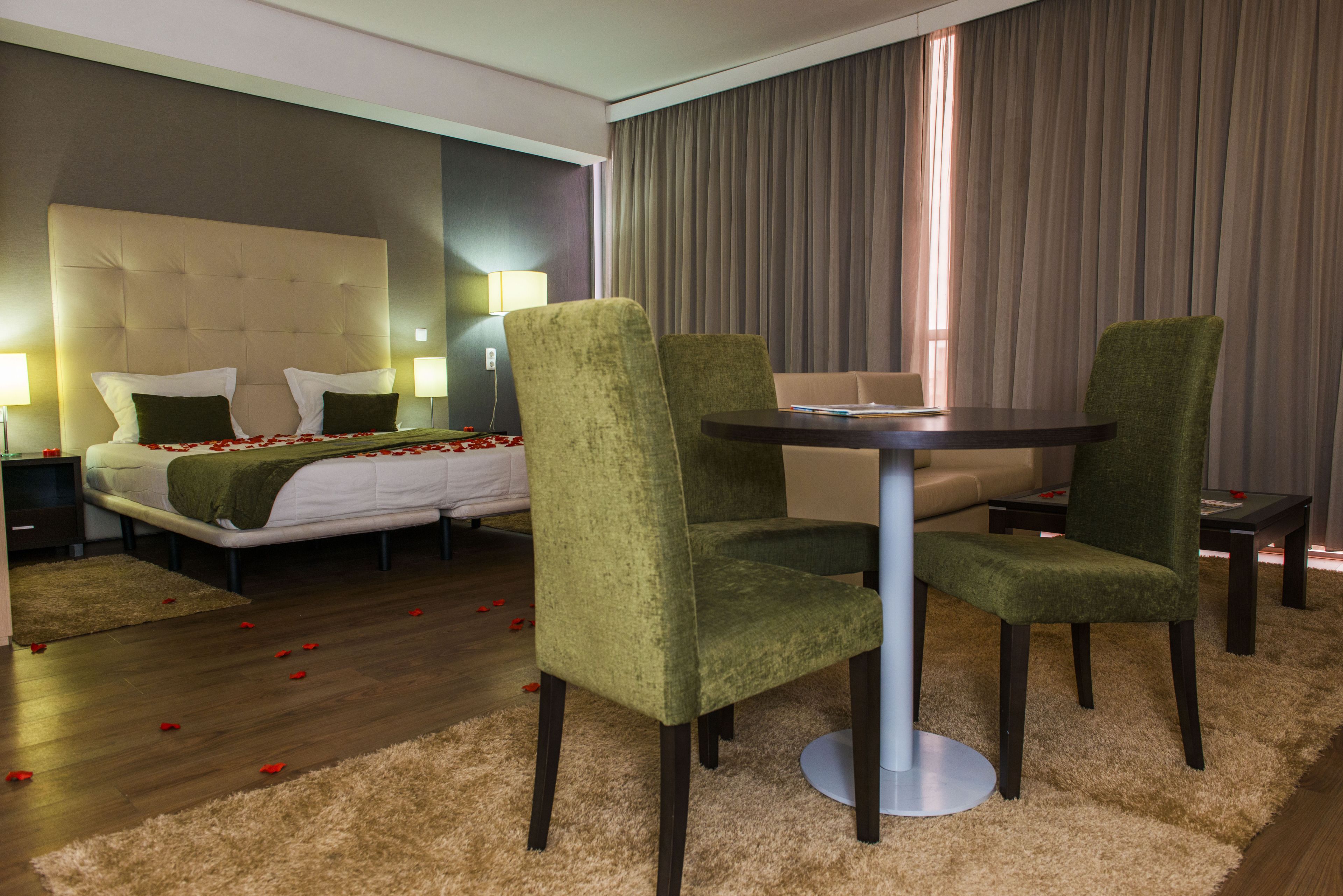 A hotel room with a bed, green upholstered chairs, a round table, and a large window with curtains.
