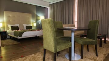 A hotel room with a bed, green upholstered chairs, a round table, and a large window with curtains.