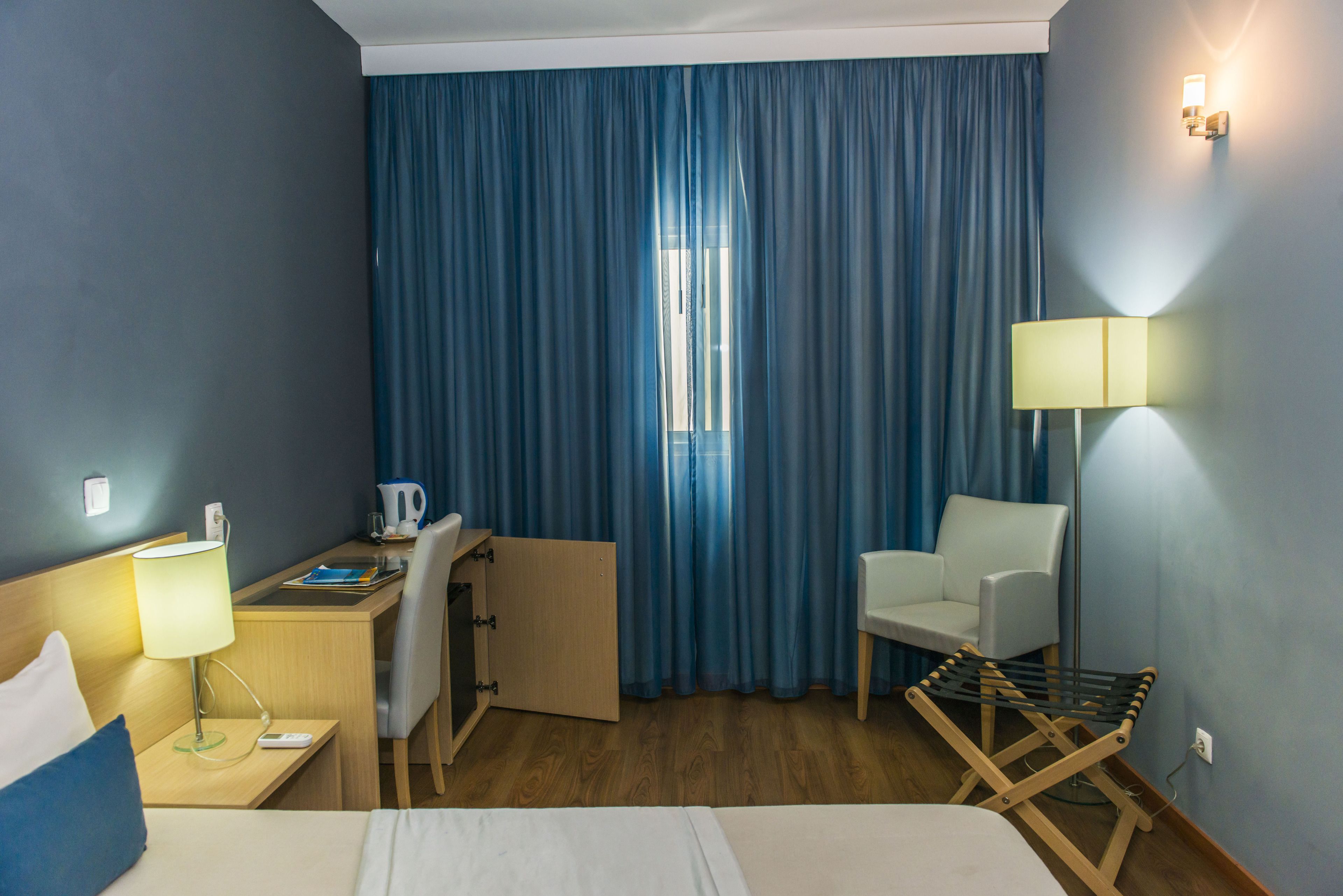 Una habitación de hotel con cama, escritorio, silla, l ámpara y ventana con cortinas.