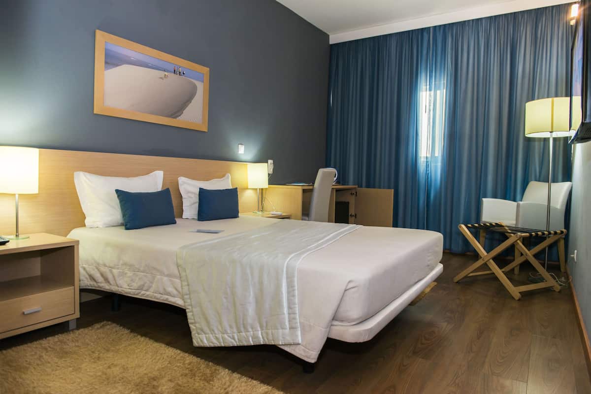 Une chambre d’hôtel avec un grand lit, deux tables de chevet avec des lampes, un bureau, une chaise et un rideau bleu.