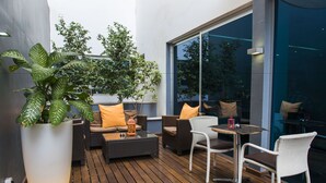 Um pátio externo com móveis de vime, uma grande planta em vaso e uma mesa de vidro.