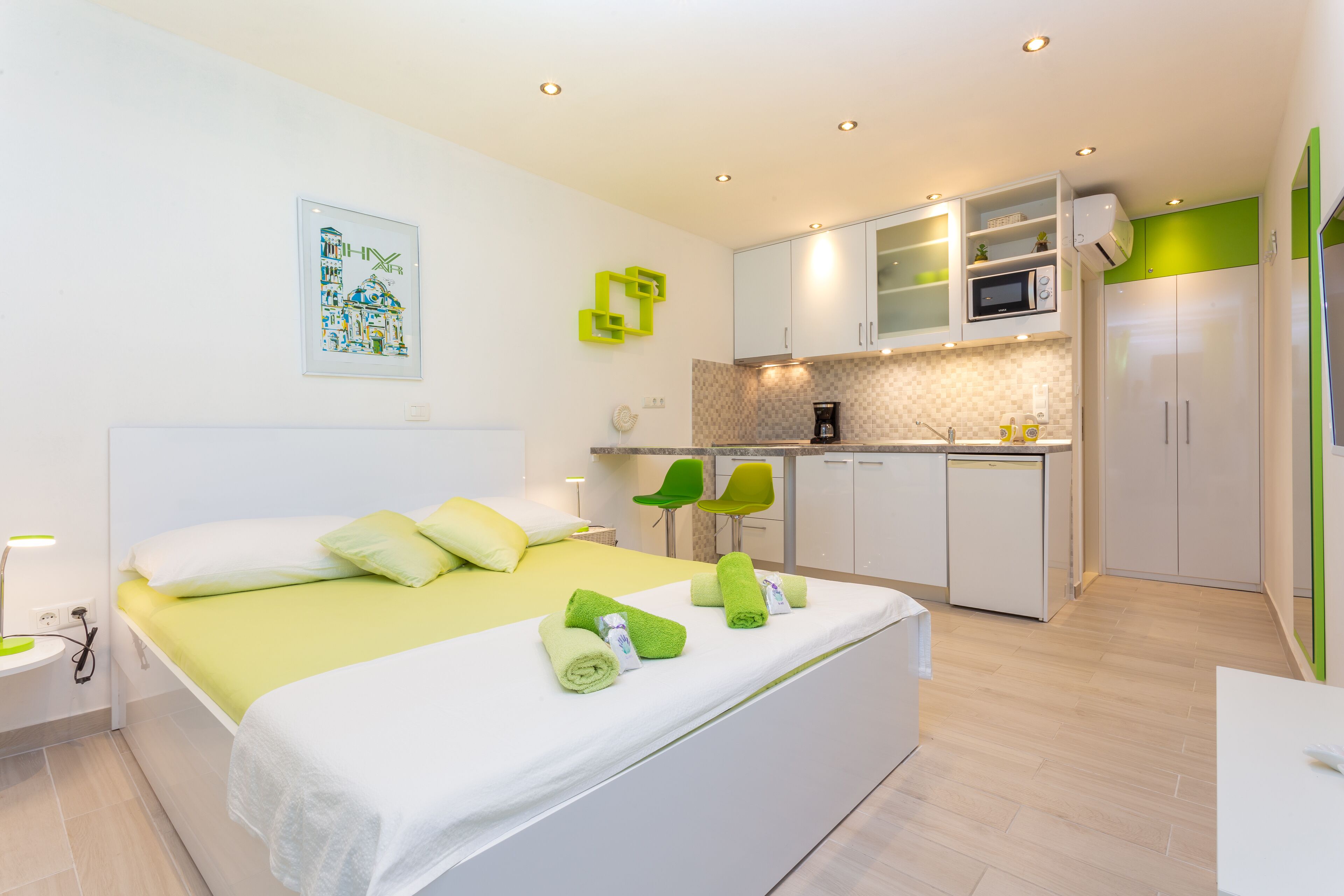 A modern hotel room with a bed, bedside table, and a kitchenette area.