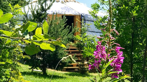 A rustic wooden cabin with a covered entrance, surrounded by lush greenery and flowering plants. - Eco Yourte Les Airelles (Saint-Antheme)