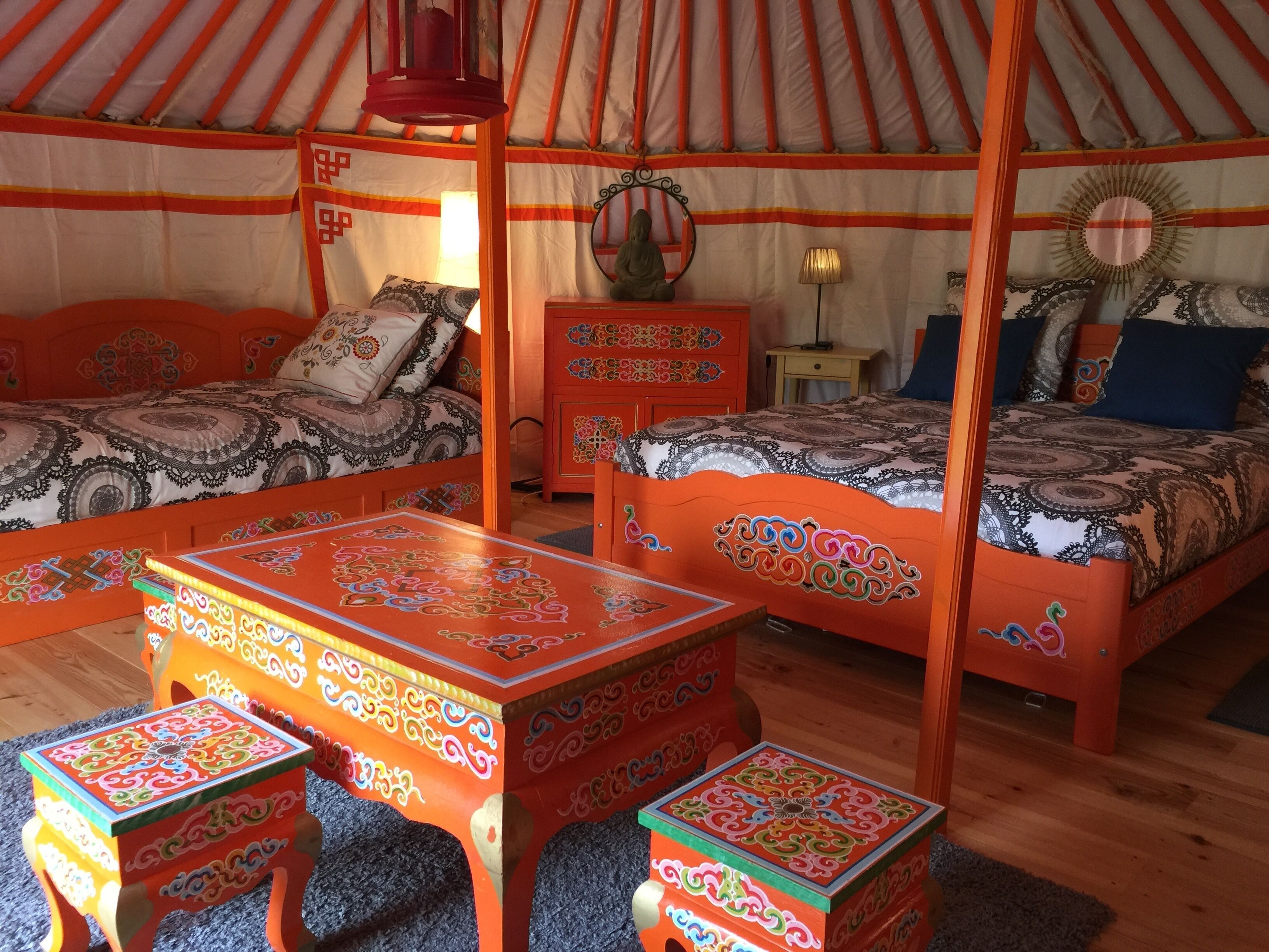 Et traditionelt yurt-interiør med en seng i midten, to mindre senge, et træbord og et dekorativt spejl.