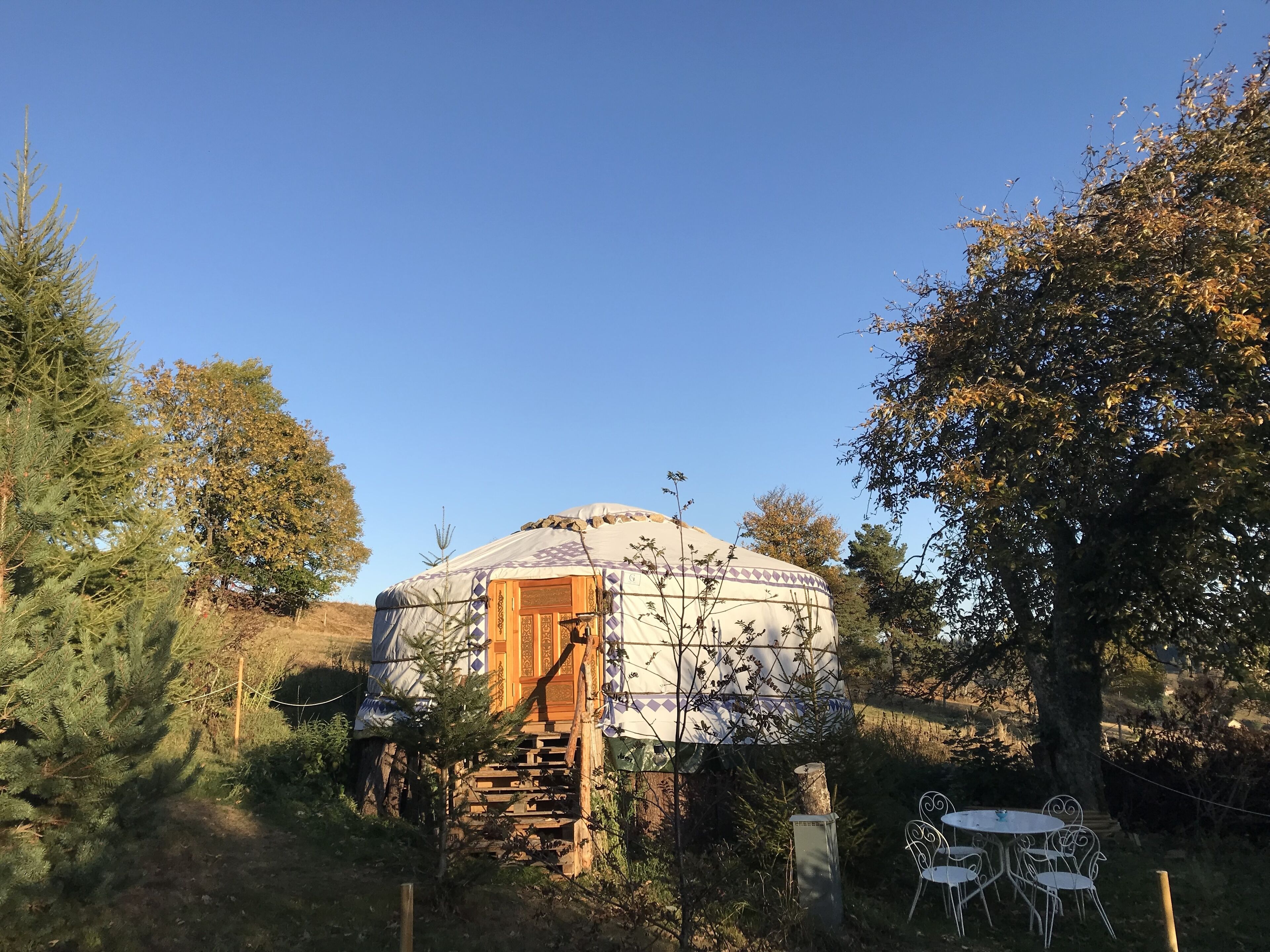 A yurt with a wooden door and a staircase leading up to it, surrounded by trees and a small outdoor seating area with metal chairs.