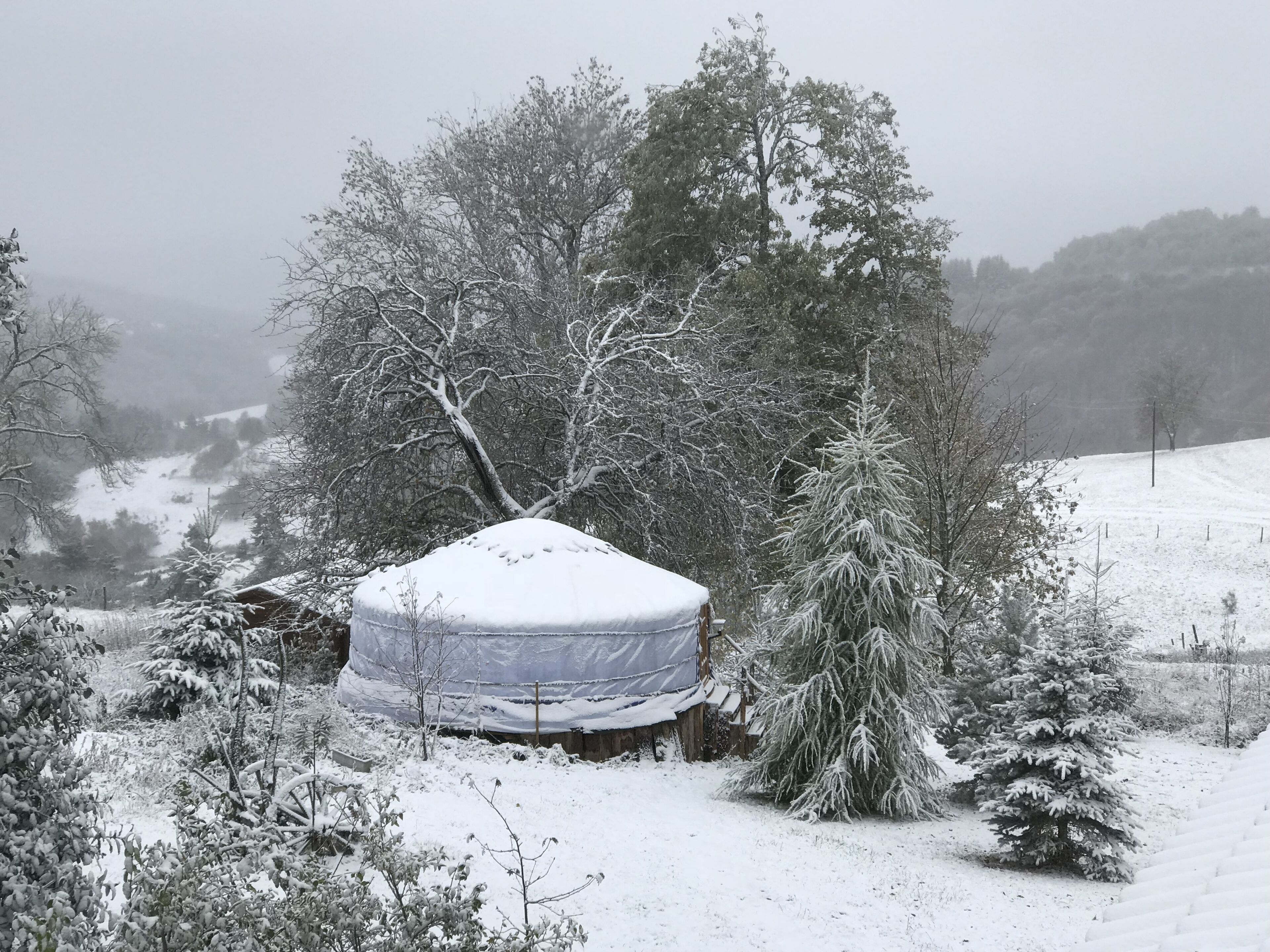 A snow-covered landscape with a tent-like structure in the foreground, surrounded by trees and a snowy hill in the background.
