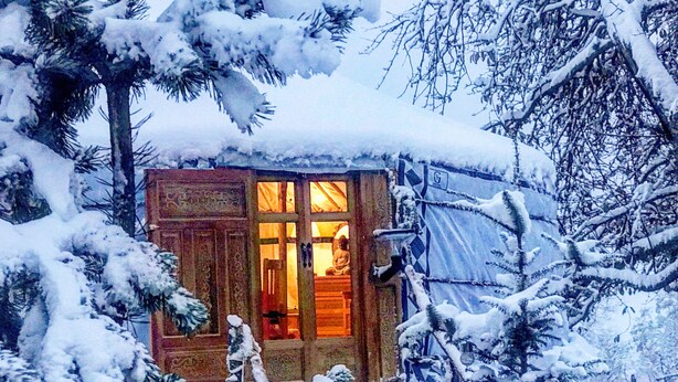 Eine kleine, mit Schnee bedeckte Hütte, durch ein Fenster ist das Innere sichtbar, umgeben von schneebedeckten Bäumen.