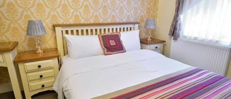 A neatly made bed with a striped blanket, a wooden headboard, and a decorative pillow. There are two bedside tables with lamps and a nightstand with a lamp. The room has patterned wallpaper and a window with curtains.