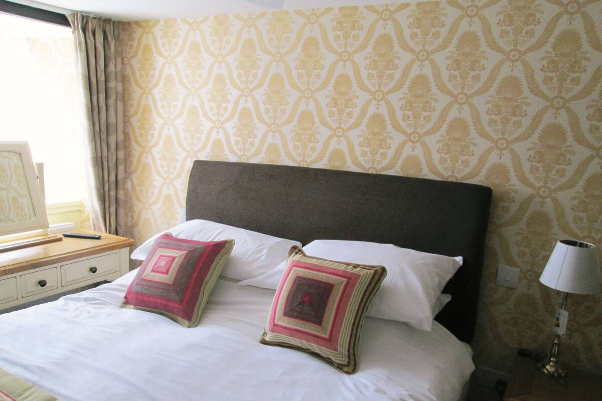 A hotel room with a large bed, two pillows, a bedside table with a lamp, and a patterned wallpaper.