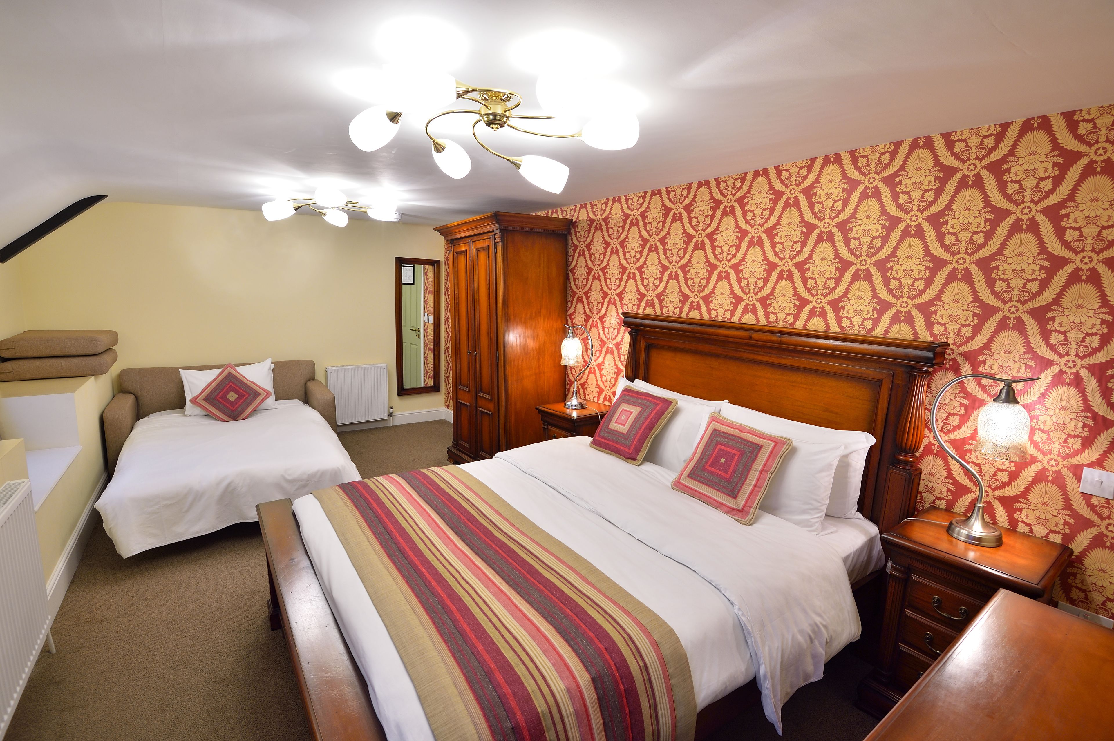Habitación de hotel con dos camas individuales, cabecera de madera, mesita de noche con lámpara y papel tapiz estampado.