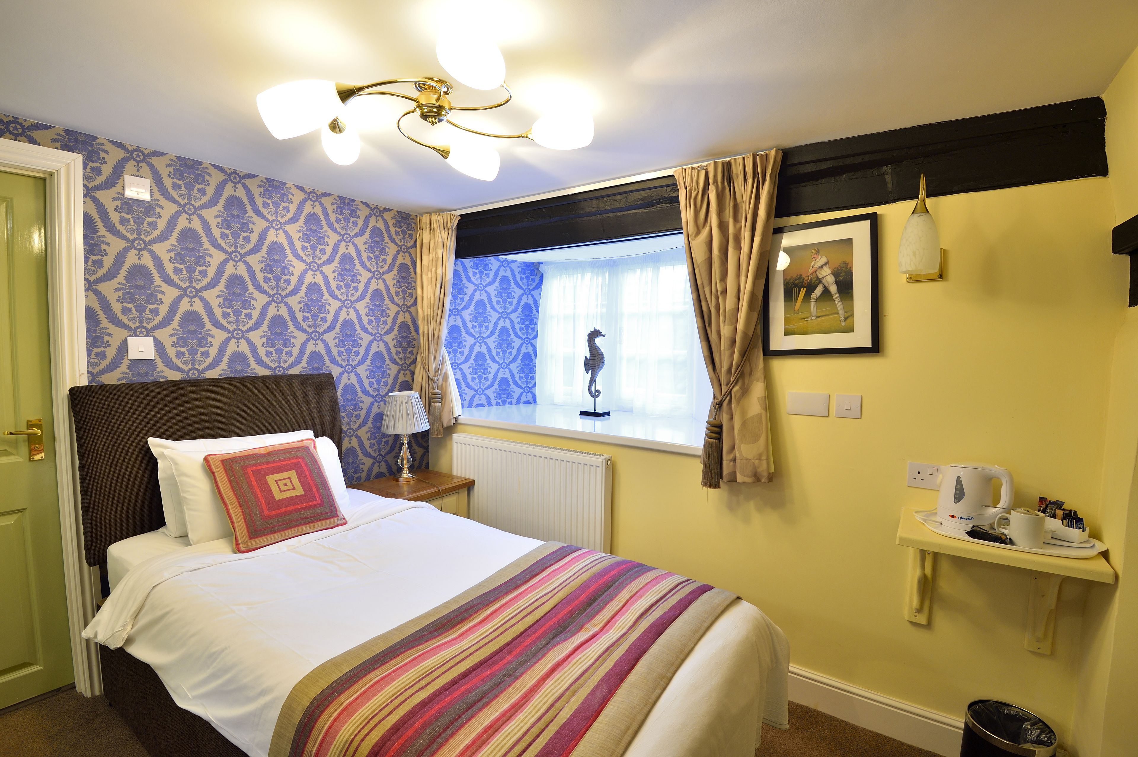 A hotel room with a bed, a window with curtains, a wall-mounted shelf with a kettle and cups, and a ceiling light fixture.