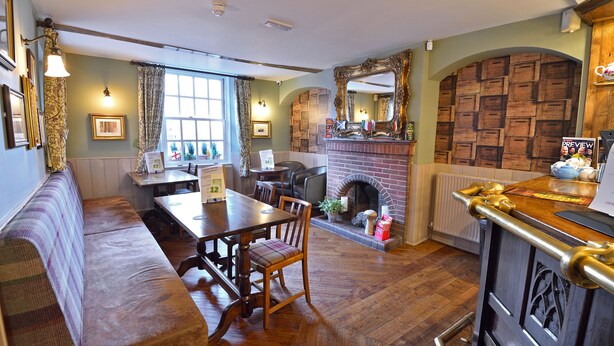 A cozy dining area with a wooden table, chairs, and a bench. There is a fireplace with a brick wall and a large mirror above it. - The Green Dragon (Wellington)
