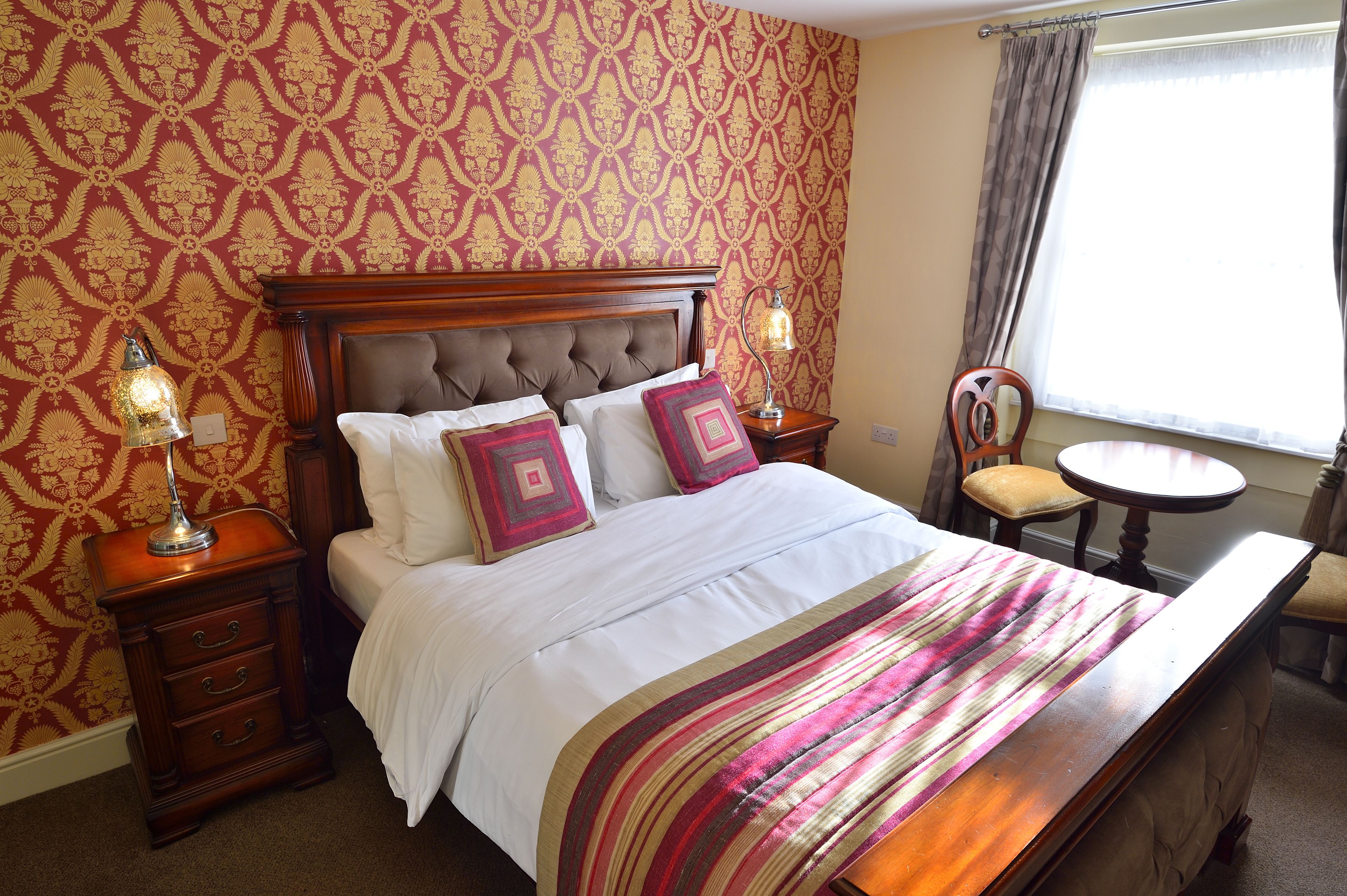 A neatly made bed with a striped bedspread, two bedside lamps, a wooden nightstand, and a chair by a window with curtains.