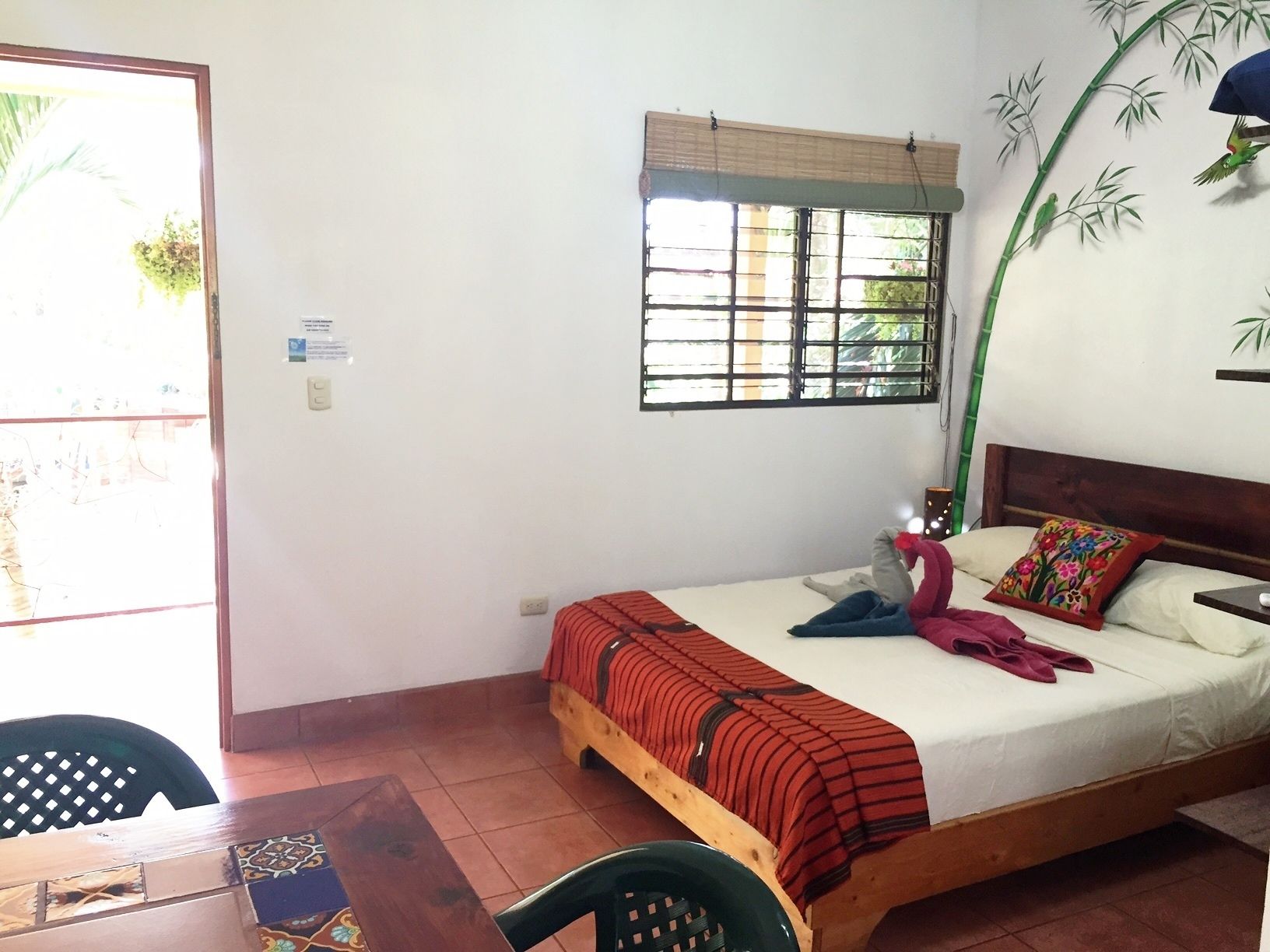 Un lit simple avec un cadre en bois, une fenêtre ornée d’un motif en bambou et une chaise avec un siège en osier tressé.