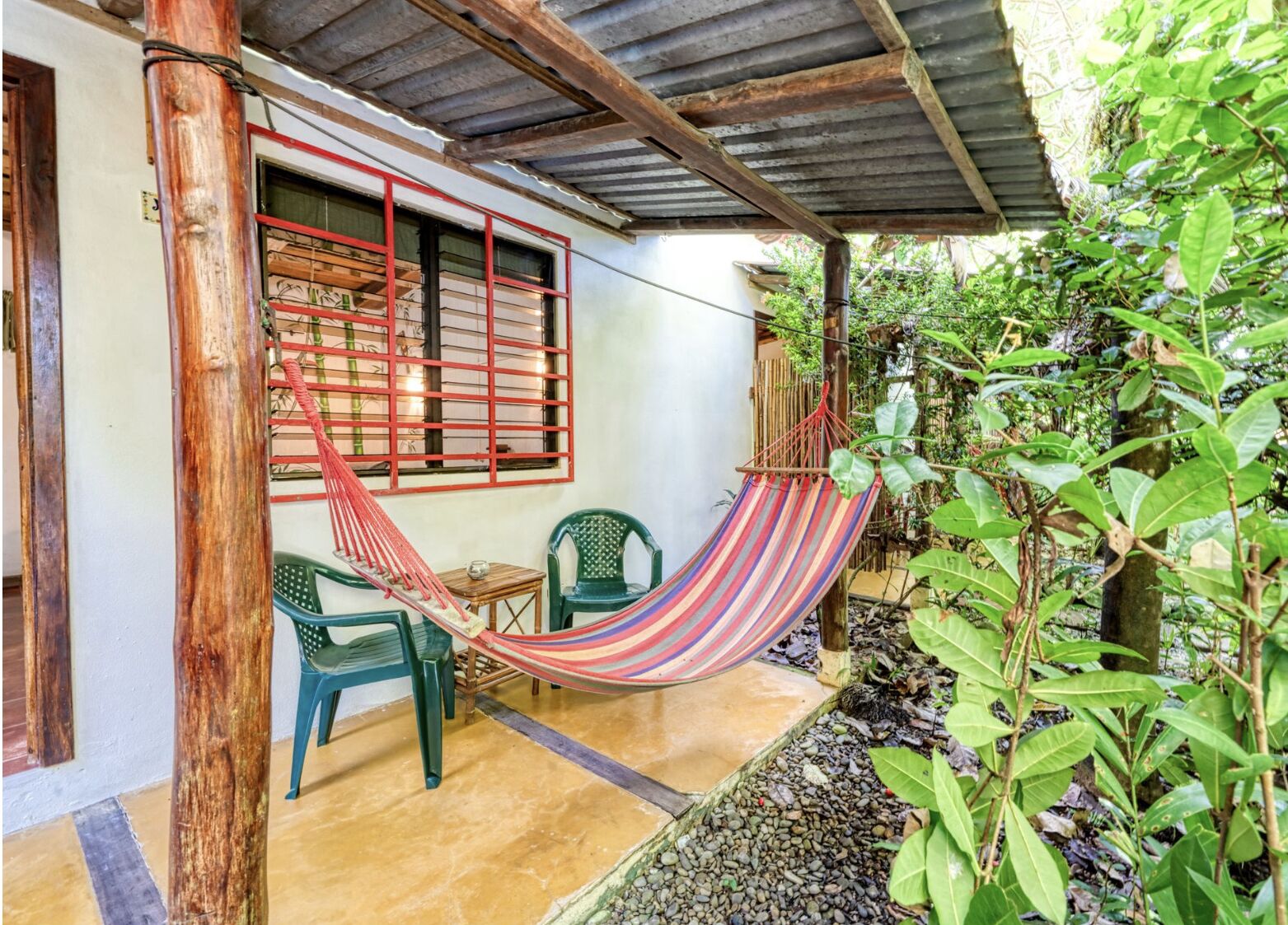 Uma rede sob um telhado de metal corrugado, uma mesa pequena e duas cadeiras em uma área externa sombreada com vista para um jardim.