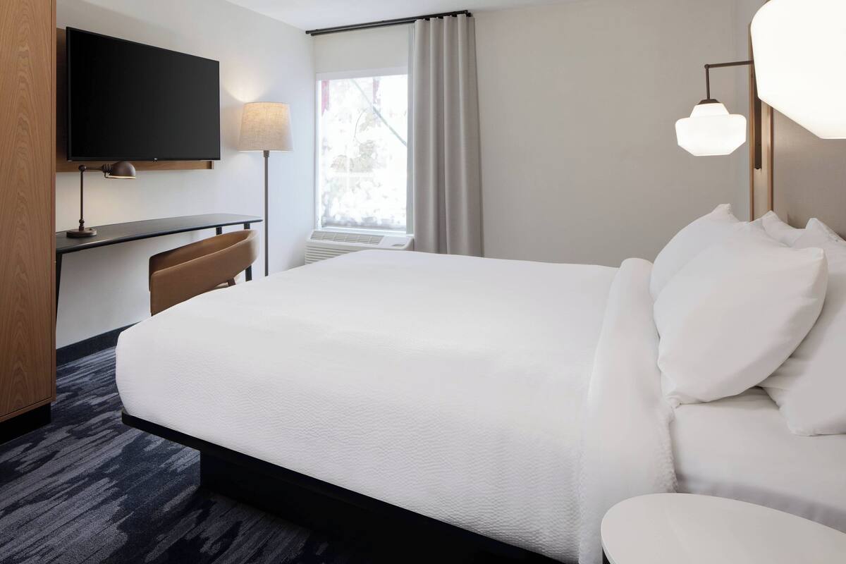 Habitación de hotel con una cama grande, un escritorio con silla, un televisor de pantalla plana montado en la pared y una ventana con cortinas.