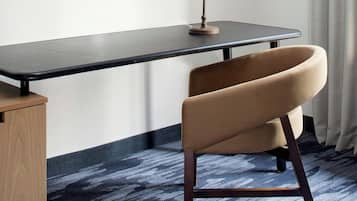 A hotel room with a flat-screen TV mounted on the wall, a desk with a lamp, a curved chair, and a patterned carpet.