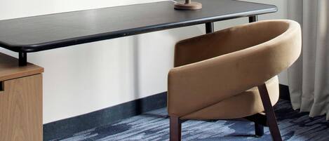 A hotel room with a flat-screen TV mounted on the wall, a desk with a lamp, a curved chair, and a patterned carpet.