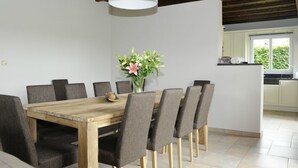 A dining area with a wooden table and grey upholstered chairs, a vase with flowers, and a kitchen in the background. - Guesthouse La Cereza (Brakel)