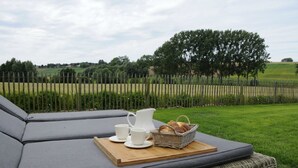 A wicker sofa with a tray of breakfast items, including bread and cups, set against a backdrop of a green field and a wooden fence. - Guesthouse La Cereza (Brakel)