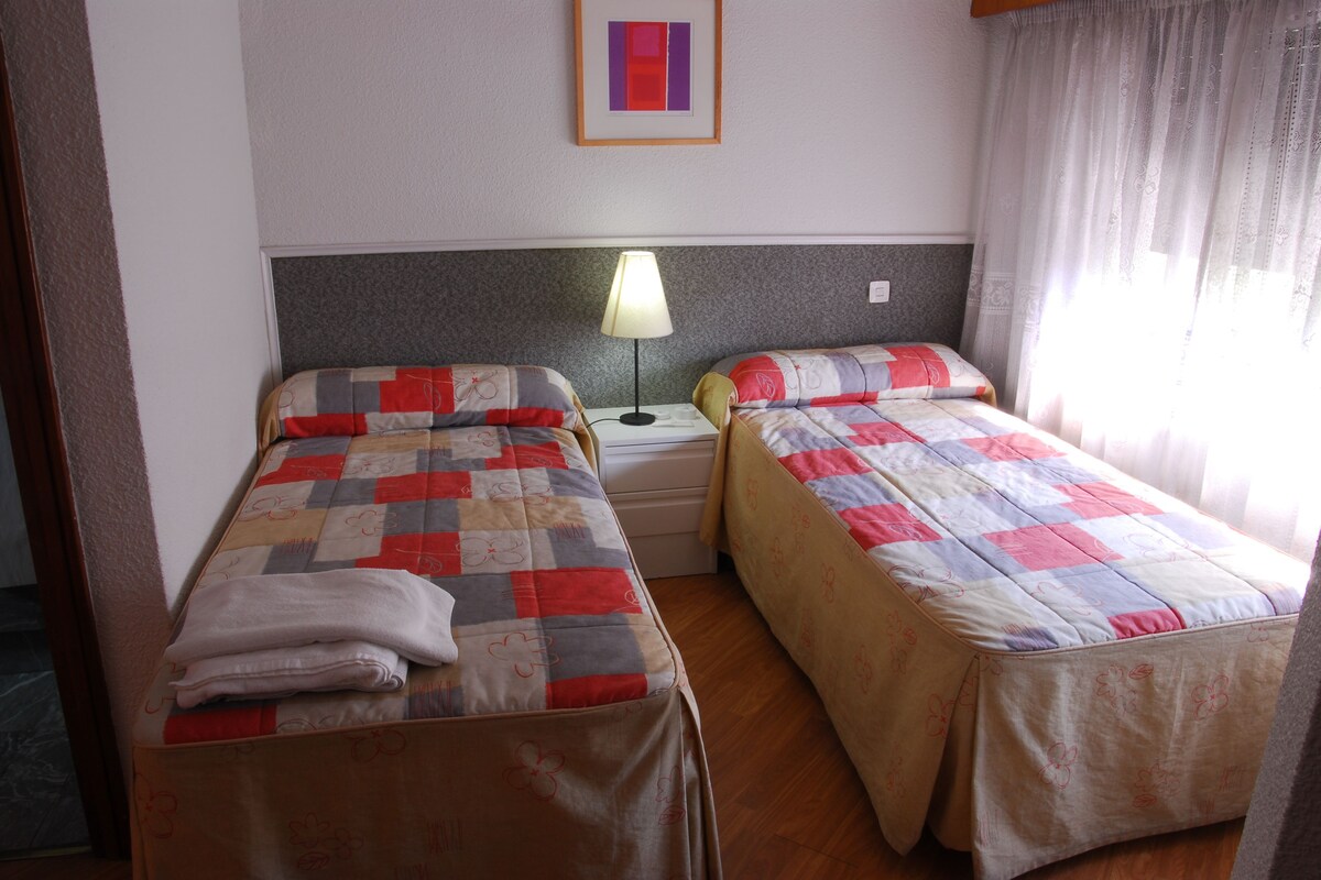 A hotel room with two single beds, a nightstand with a lamp, a framed picture on the wall, and a window with curtains.