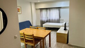 A compact hotel room with a wooden dining table, two chairs, a small sofa, a bed with a white mattress and a TV on a stand. - Apartamentos Goya 75 (Madrid)