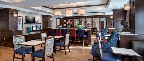 A spacious hotel lobby with a central bar area, seating arrangements, and a television mounted on the wall.