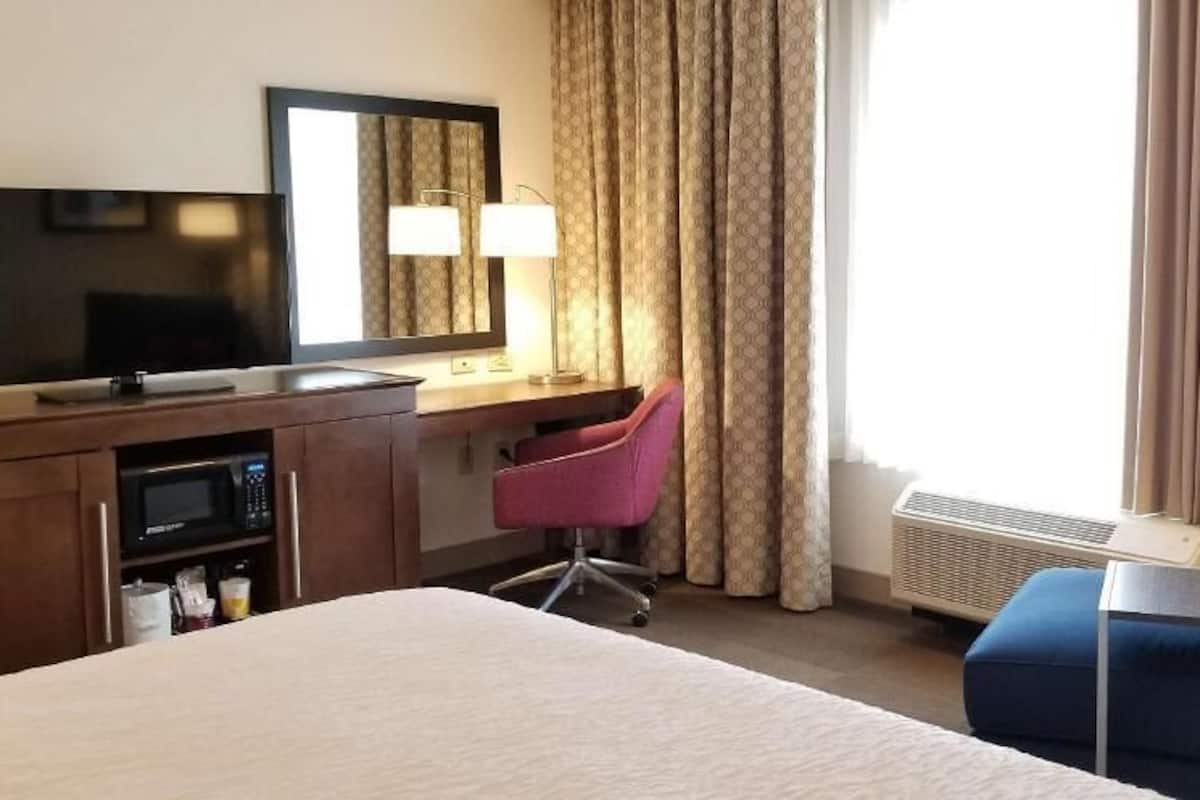 A hotel room with a bed, a desk with a TV, a chair, and a window with curtains.