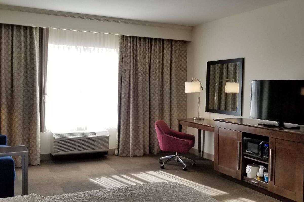 A hotel room with a blue armchair, a red office chair, a desk with a television, and a large window with curtains.