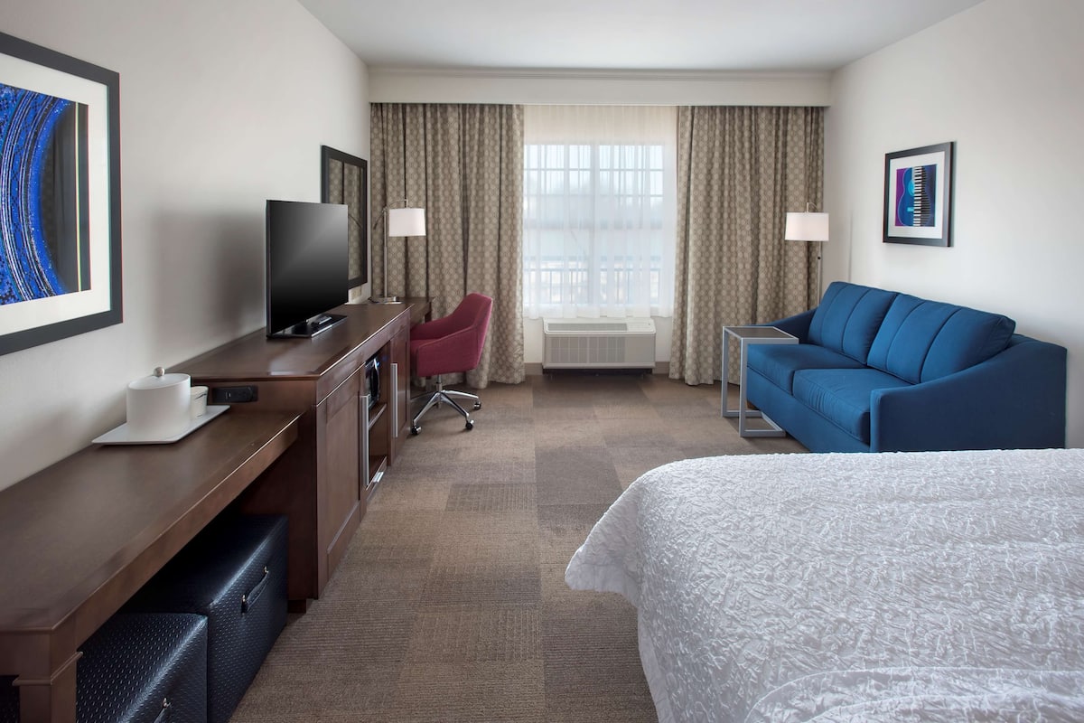 A hotel room with a bed, a desk with a TV, a blue sofa, and a chair.
