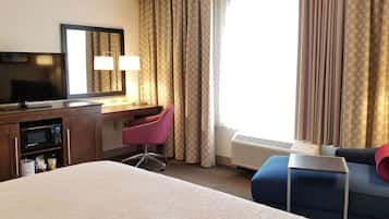 Habitación de hotel con cama, escritorio con computadora, televisión, silla y ventana con cortinas.