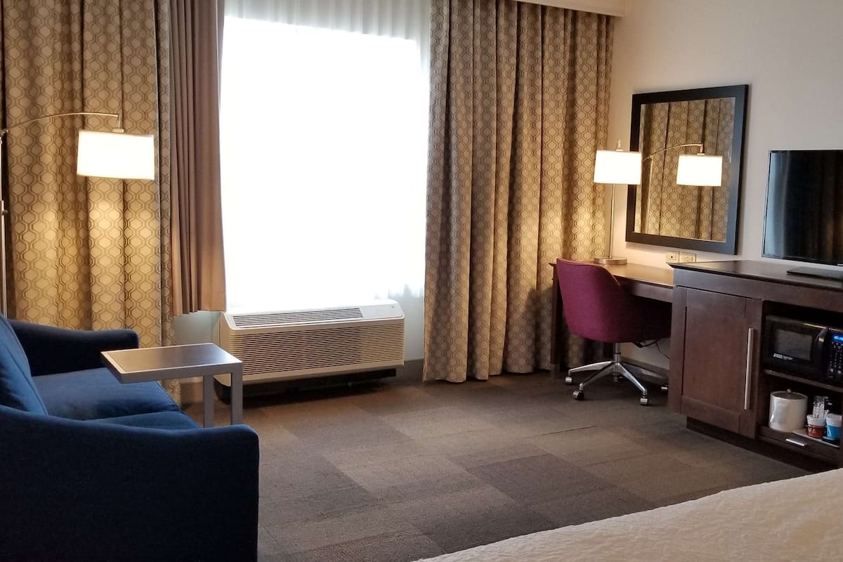 A hotel room with a bed, a desk with a chair, a television, a mirror, and an air conditioning unit.