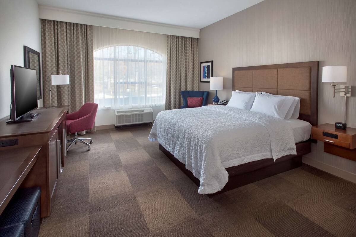 A hotel room with a bed, a desk with a TV, a chair, and a window with curtains.