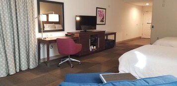 A hotel room with a bed, a desk with a computer, a chair, a TV, and a bathroom door.